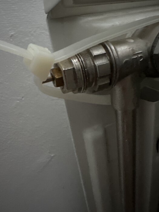 Närbild av en radiatorventil med en slangklämma runt anslutningen, monterad på en vägg mot en grå bakgrund.