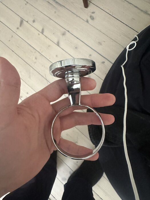 Kromat badrumstillbehör i handen, potentiellt handdukshållare från Miller, med trägolv i bakgrunden.