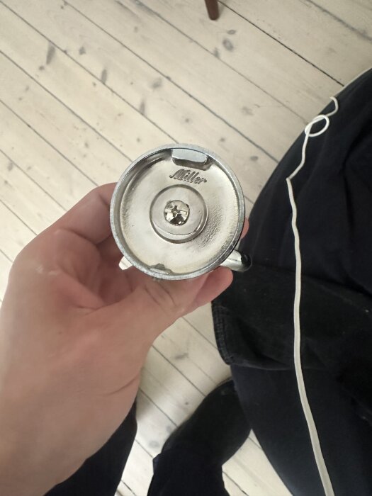 Hand håller i en rund, silverfärgad Miller-badrumstillbehör med skruv i mitten, oklart hur det ska monteras.