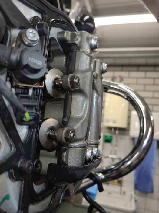 Motorcykelmotor med ventiljusteringsdelar synliga, inga shims. Verkstadsmiljö med tegelvägg i bakgrunden.
