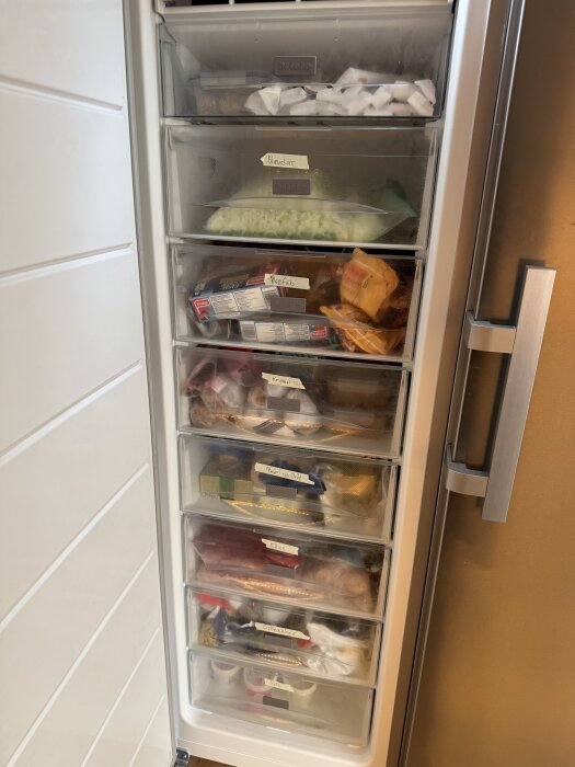 Öppen frys med flera lådor fyllda med matvaror, däribland hackad lök och andra ingredienser, märkt med etiketter för organisering.