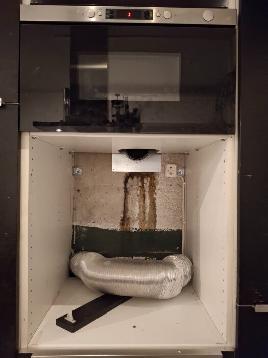 Ventilationshål och rör bakom inbyggd mikrovågsugn, med synlig köksvägg och eluttag i skåp mellan mikrovågsugn och ugn.