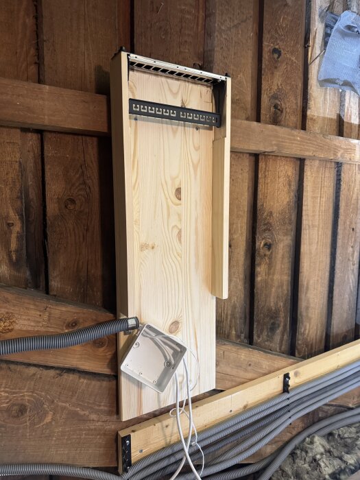 Hemmabyggt nätverksrack med träpanel och slangar fästa med klämmor mot en trävägg i ett hus, med utrymme för framtida switchinstallation.