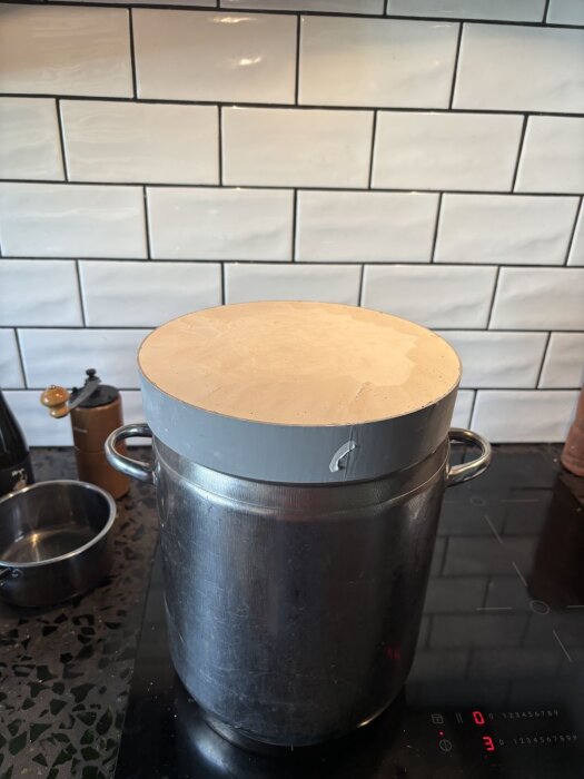 Gryta med lock av en form för glasgjutning, ståendes på en svart spishäll, med kakelvägg i bakgrunden.