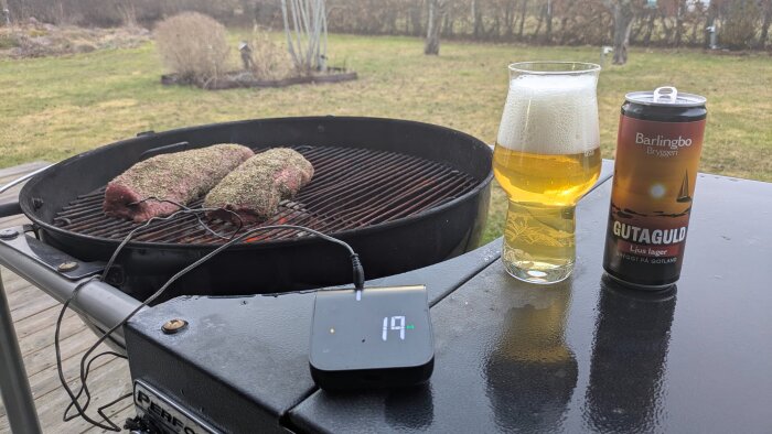 Grillad köttbit med termometer på utegrill, ölglas och burk bredvid, i en trädgårdsmiljö.