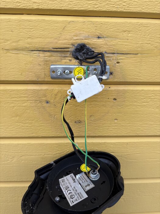 Öppen utomhusarmatur med jordkablar kopplade till fästet och elcentral, mot gul trävägg. Lampans hölje och sladdanslutningar synliga.