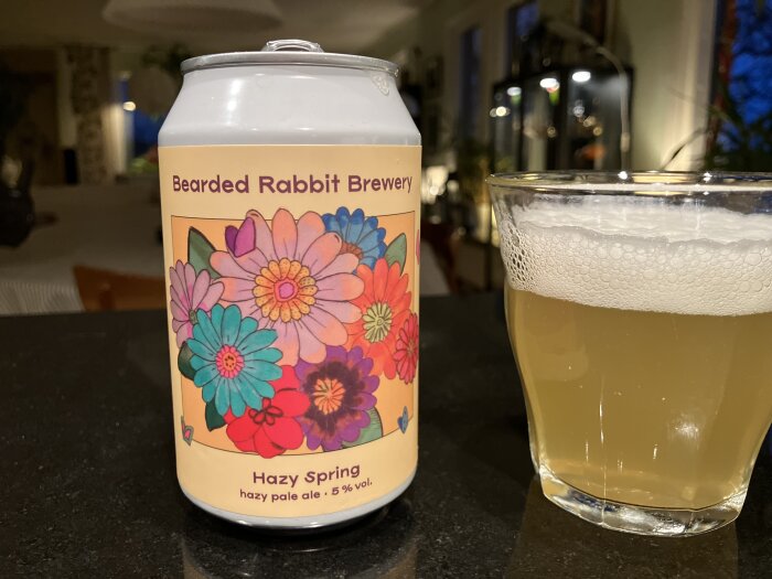 En burk Bearded Rabbit Brewery Hazy Spring ale med blommig etikett bredvid ett glas med skummande ljus öl, inomhusmiljö i bakgrunden.