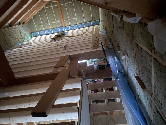 Trätrappa i nybyggt hus, med 3 steg på plats. Byggmaterial ligger ovanpå vilplanet. Isolering och träkonstruktion syns på väggarna.