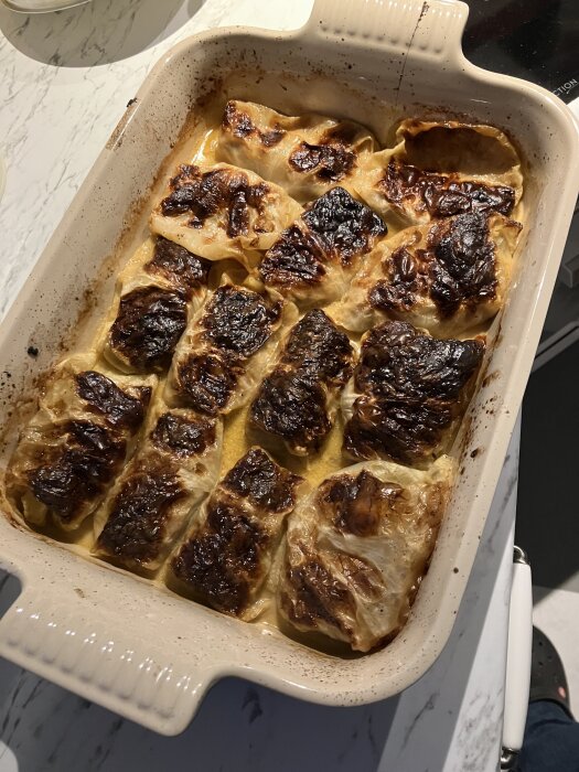 Ugnsform med flera kåldolmar, gyllenbruna och gratinerade, på ett marmorbord.