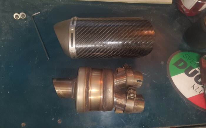 Ljuddämpare med kolfiberhölje och metallkomponenter ligger på ett bord bredvid verktyg och en Ducati-logga.