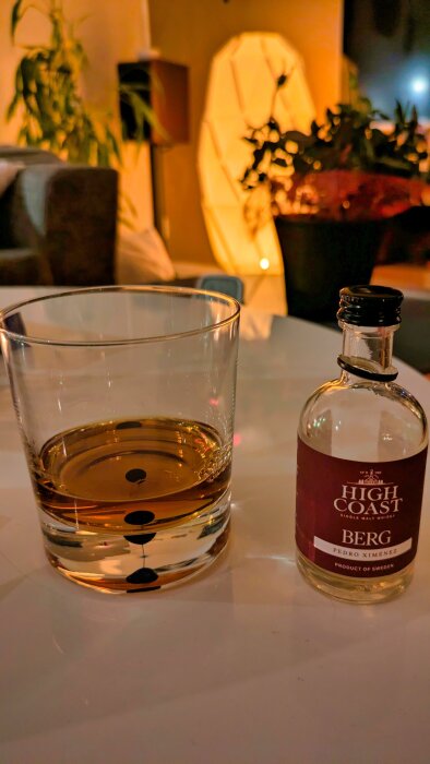 Glas med whisky och en flaska High Coast Berg whisky står på ett bord i ett mysigt upplyst rum med växter i bakgrunden.