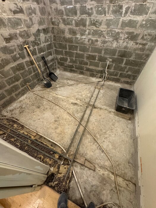 Bild av ett källarrum under renovering, med ojämn betongplatta, synliga rör och väggar av block med redskap lutade mot dem.