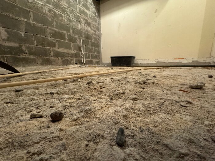 Ojämn betongplatta i en källare med små hål och sprickor, delar av en vägg syns med rå yta och oklara material, rör på golvet.