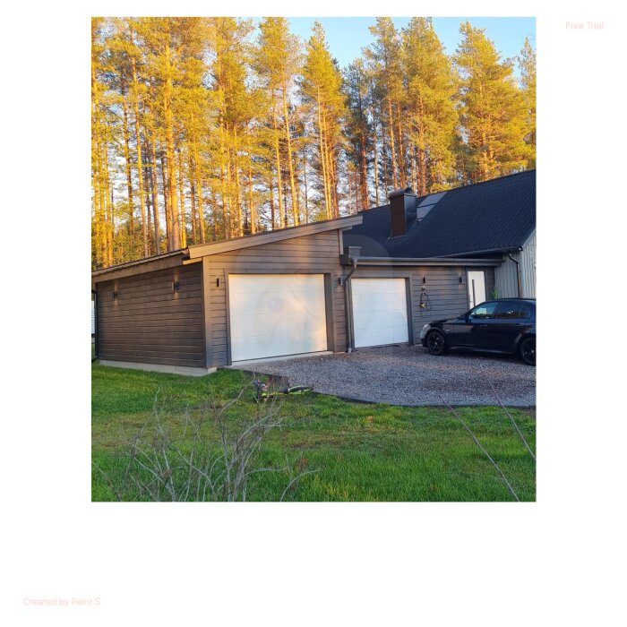 Grå garagebyggnad med två stora vita portar, grusad uppfart, och en svart bil parkerad framför, omgiven av skog.