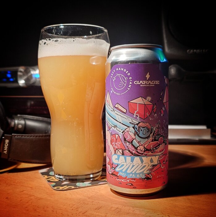 Ett glas med oklar, ljusgul dryck bredvid en färgglad burk märkt "Galaxy Divide" på ett träbord.