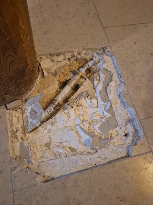 Träpelare på kakelgolv där en bit är uppbruten för att exponera ett rör i undergolvet, omgivet av sprucket cement.