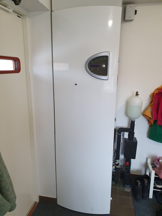 Vit bergvärmepump installerad inomhus bredvid dörr, med rör och vattenbehållare intill.