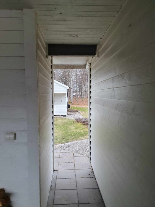 Smal gång med vita träväggar och tak; leder ut mot en gräsyta och ett vitt hus i bakgrunden. Stengolv med grå plattor.