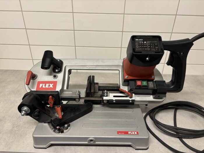 Metallbandsåg FLEX SBG4910 med röd och svart design på ett bord framför en kaklad vägg, inklusive strömbrytare och anslutningskabel.