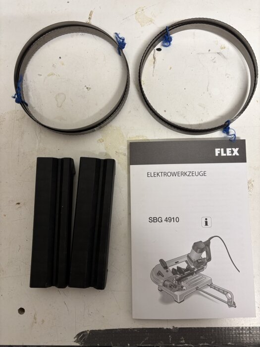 Två extra bandsågblad, rörbackar och en instruktionsbok för FLEX SBG4910 metallbandsåg på ett bord.
