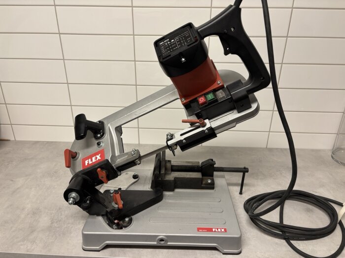 Metallbandsåg FLEX SBG4910 på ett bord med sladd och skruvstäd. Sågen är i gott skick, används för hobby.