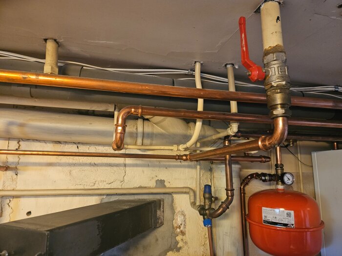 Rörinstallation i ett hus med kopparrör, ventiler och en expansionskärl märkt "Varem". Beige målade väggar och tak syns i bakgrunden.