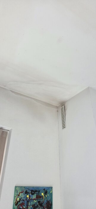 Missfärgat tak och vägg nära ventilationsgaller, antydan till sot eller fläckar, ovanför en färgglad tavla.
