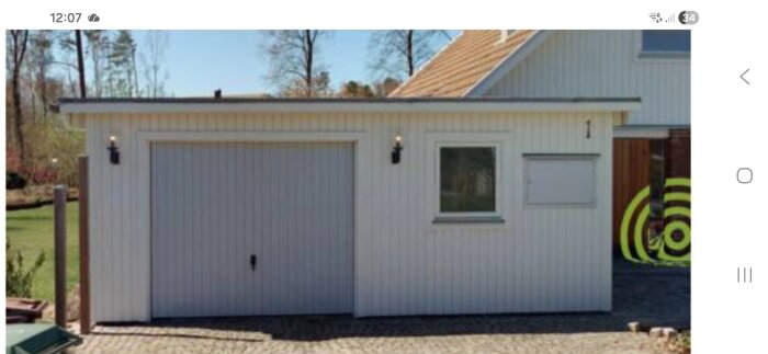 Vit träfasad på garage med låglutande tak, garageport, fönster, lampor och stenlagd uppfart under blå himmel.