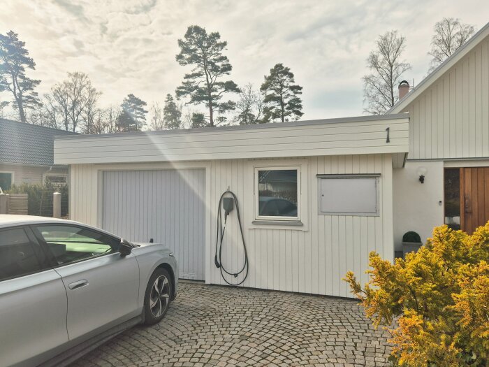En vit garagebyggnad med låglutande tak, elbilsladdare och en parkerad silverfärgad bil på stenlagd uppfart framför ett hus.