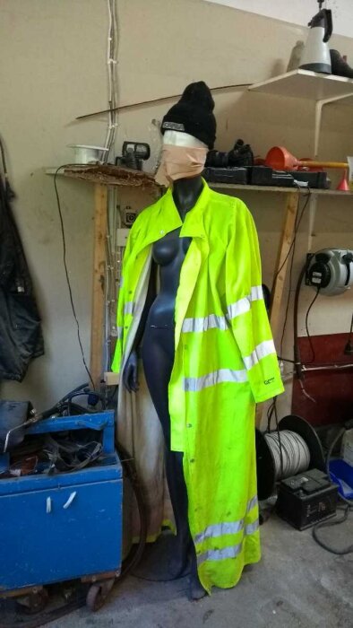 Mannequin i gul varseljacka och mössa, stående bredvid vägg med dåligt klamrade kablar, verktyg och hyllor i bakgrunden.