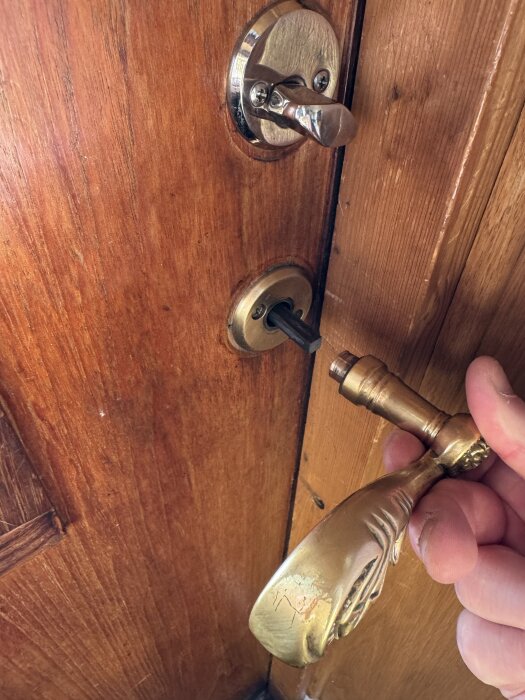 Guldhandtag på träfärgad dörr är löst och hålls i handen. Det saknas en fästskruv för att stabilisera handtaget på insidan.