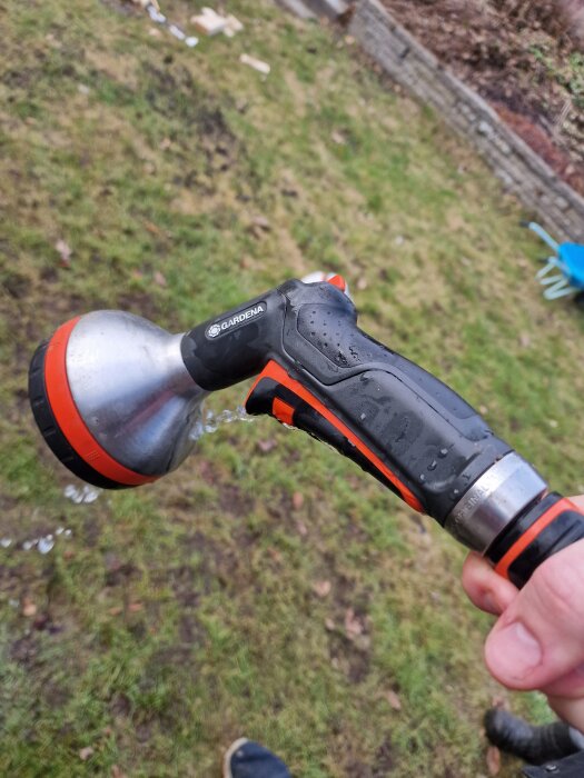 Läckande munstycke från Gardena med vattenslang i trädgård. Vatten strömmar ut från sida. Användarens hand håller munstycket.