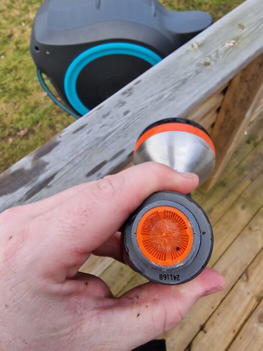 Hand som håller en orange och grå trädgårdsslanganslutning nära en träräcke, med en rulle trädgårdsslang i bakgrunden.
