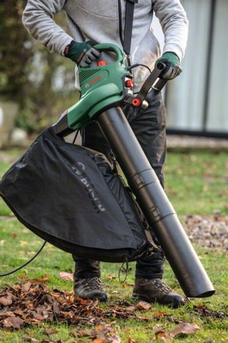 En person använder en grön lövblås med en svart uppsamlingspåse på en gräsmatta med höstlöv.