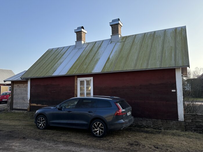 Rött hus med grönt tak och två skorstenar. Skorstensområdet är rent från mossa, medan resten av taket har grön beläggning. Grå bil parkerad framför.