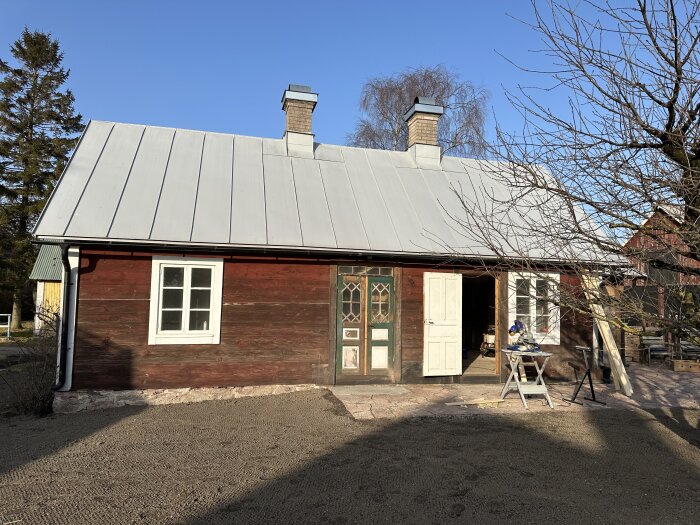 Rött timmerhus med vit dörr och fönster, plåttak och två skorstenar. Verktyg och brädor syns framför huset, omgivna av träd och klarblå himmel.
