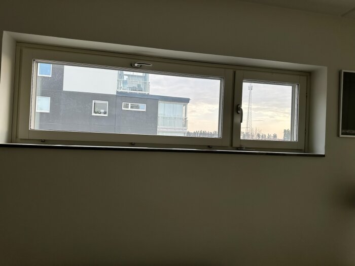 Utsikt genom ett fönster med vit karm visar en modern byggnad och en telemast i bakgrunden under en molnig himmel.
