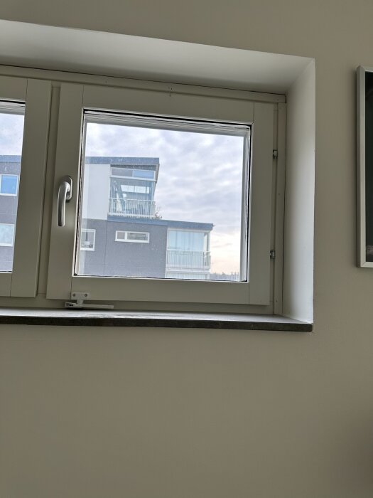 Vy av ett fönster inifrån som visar en del av en byggnad utanför under en molnig himmel.
