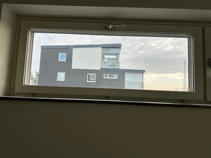 Utsikt genom fönster mot en modern byggnad med grå fasad och glasdetaljer, himlen är molnig med ljus i horisonten.