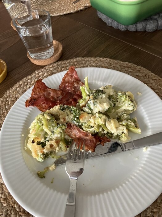 Broccoligratäng med stekt lök, keso, västerbottenost och bacon på en tallrik, serverad med bestick och ett glas vatten.