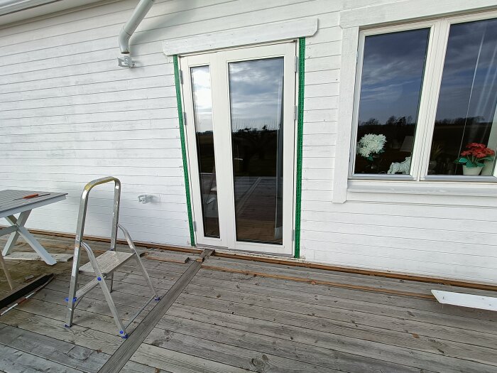 Vit altandörr med fuktproblem, trädäck skruvat mot huspanel; stege står intill. Problem med fukt vid tröskel och smyg av MDF.