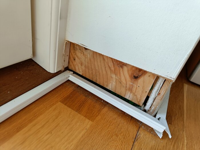 Närbild på en nedre hörna av en altandörr med en skadad MDF-panel och synlig träregel, omgiven av trägolv och vitmålad kant.