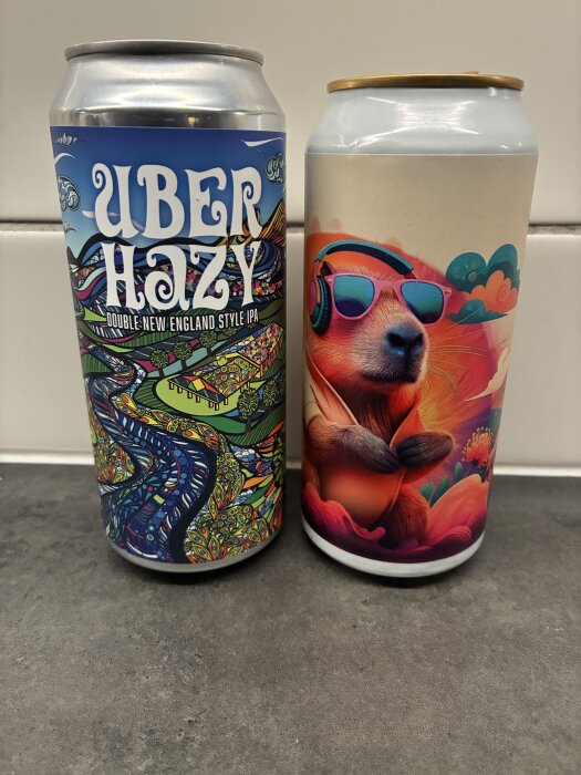 Två färgglada ölburkar på ett grått bord, en med "Uber Hazy" etikett och en med illustration av ett djur med hörlurar och glasögon.