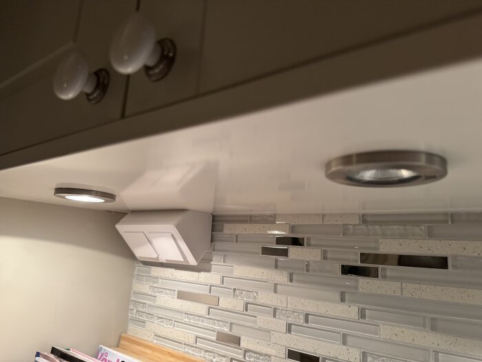 Skåpluckor med två spotlights monterade under, strömbrytardosa på väggen och mosaikplattor som stänkskydd i kök.