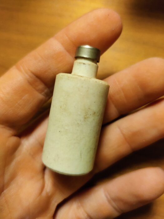 En hand håller en liten, cylindrisk keramik- eller porslinsdel med en metalltopp, eventuellt en del av en byggkomponent.