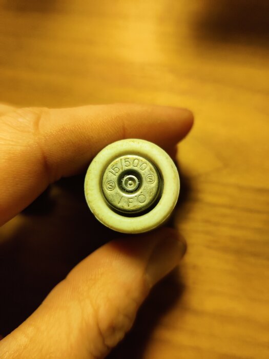 Närbild på en persons hand som håller en liten elektrisk komponent med märkningen "15/500" och "YFO" mot en träbakgrund.
