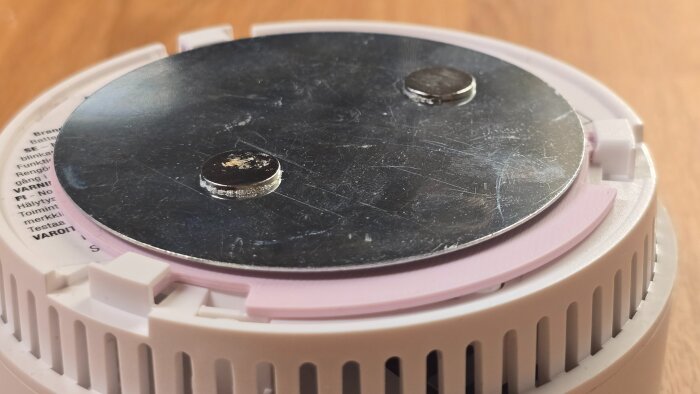 Rund vit brandvarnare med monterad metallplatta och två magneter för magnetfästen, visas ovanifrån på ett bord.
