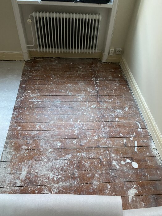 Gammalt trägolv med spår av fernissa, färg- och spackelfläckar, delvis exponerat intill en vit radiator och vägguttag.