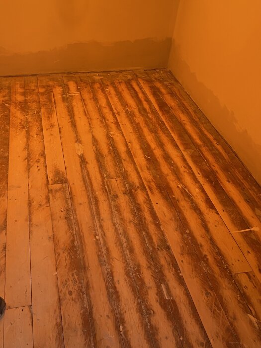 Gammalt trägolv av furu eller gran med spikmärken, fernissabehandlat med färgfläckar, i behov av renovering eller parkettbyte.