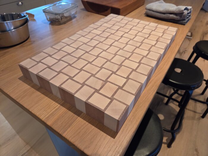 Träskärbräda med rutmönster ligger på en köksbänk i trä, omgiven av hushållsföremål såsom en metallskål och vikta kökshanddukar.